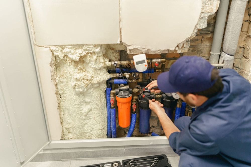 Plumbing by Jake provides commercial water treatment and water softener installation services in Golden Valley, Arizona for homes and businesses dealing with hard-water buildup and plumbing system issues. This image shows a professional plumbing technician servicing a water treatment and filtration system installed behind an open wall utility access panel with visible filtration canisters, shutoff valves, plumbing connections, and blue water supply lines. The organized plumbing setup highlights professional installation and repair standards used to improve water quality, reduce mineral buildup, and help protect plumbing systems in Arizona properties.