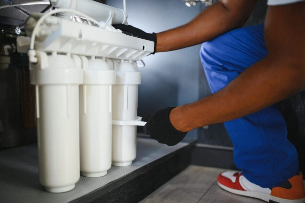 A plumbing technician services a commercial water treatment and softener installation system beneath a sink in Kingman, Arizona while replacing reverse osmosis water filters. The image shows connected filtration canisters, water purification tubing, and a professionally maintained water treatment system designed to improve water quality and reduce hard water minerals in residential and commercial plumbing systems. Plumbing by Jake provides commercial water treatment and softener installation services in Kingman AZ including reverse osmosis system maintenance, water filtration upgrades, and hard water treatment solutions. Customers in Kingman can contact Plumbing by Jake at 928-615-8228 for water softener installation, filter replacement, and commercial water purification services.