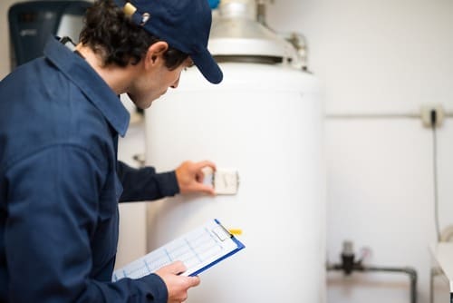 A plumbing technician performs a tankless water heater installation in Fort Mohave, AZ while inspecting system controls and water connections inside a residential utility room. Plumbing by Jake provides tankless water heater installation services for homeowners who need dependable hot water and energy-efficient equipment. The image shows a technician reviewing installation details near a water heating system with visible plumbing connections and utility components. Homeowners in Fort Mohave can contact Plumbing by Jake at (928) 615-8228 for tankless water heater installation, replacement, and hot water system services.