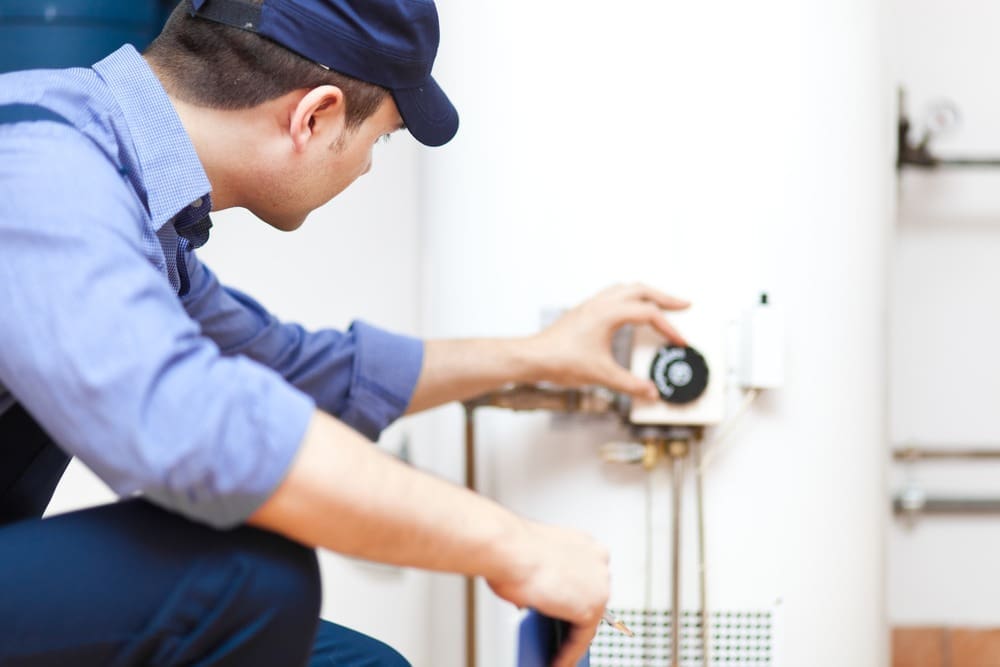 A licensed plumbing technician adjusts the temperature control on a newly installed residential water heater during a water heater installation and replacement service in Mohave Valley, AZ. The image shows a tall tank-style water heater with visible plumbing connections, water supply lines, shut-off valves, and utility room components commonly found in Arizona homes. Plumbing by Jake provides water heater installation and replacement services in Mohave Valley, Arizona, helping homeowners maintain reliable hot water systems.
